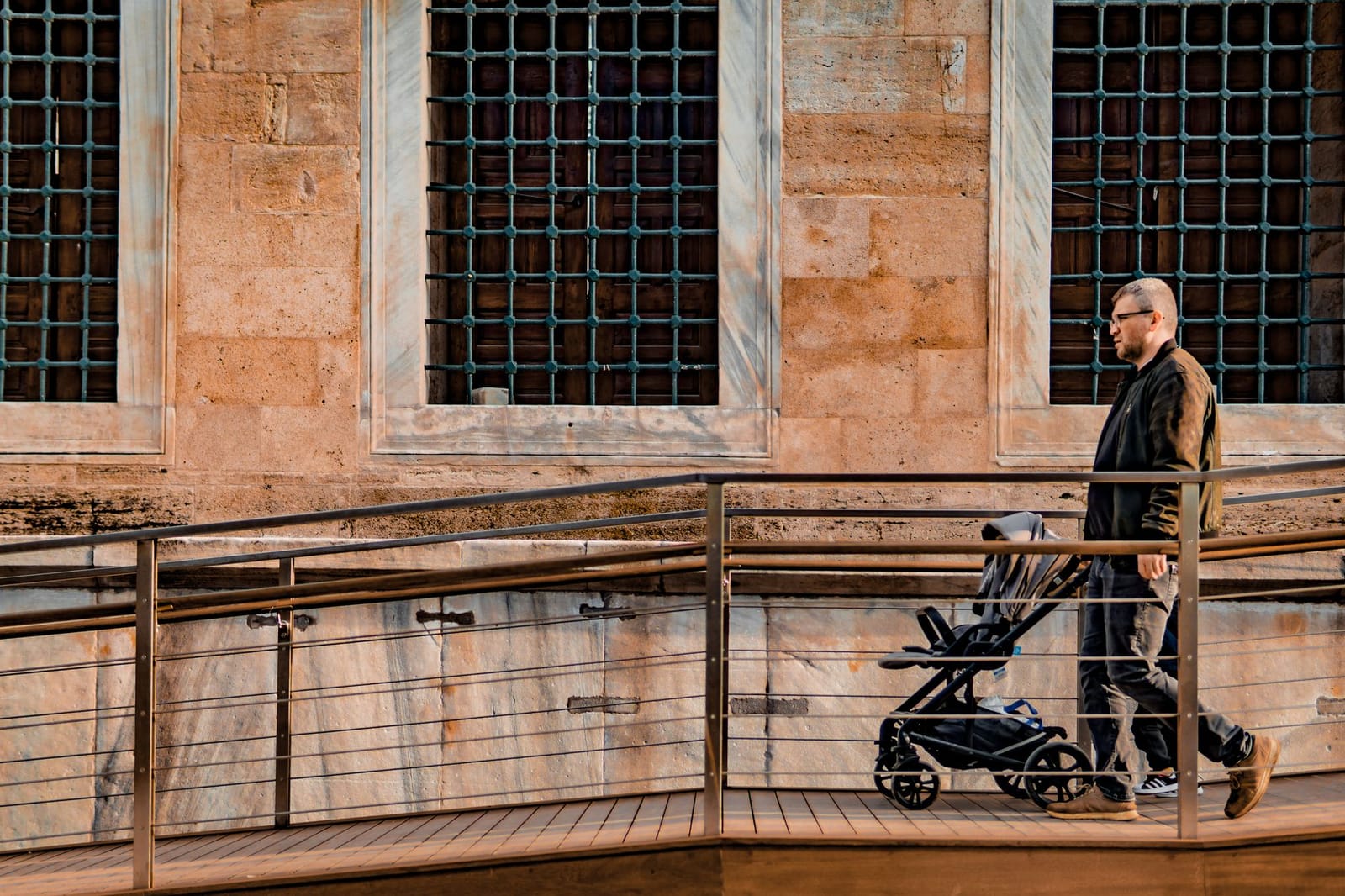 A man pushes a stroller up an accessible wooden ramp beside an ancient stone building.