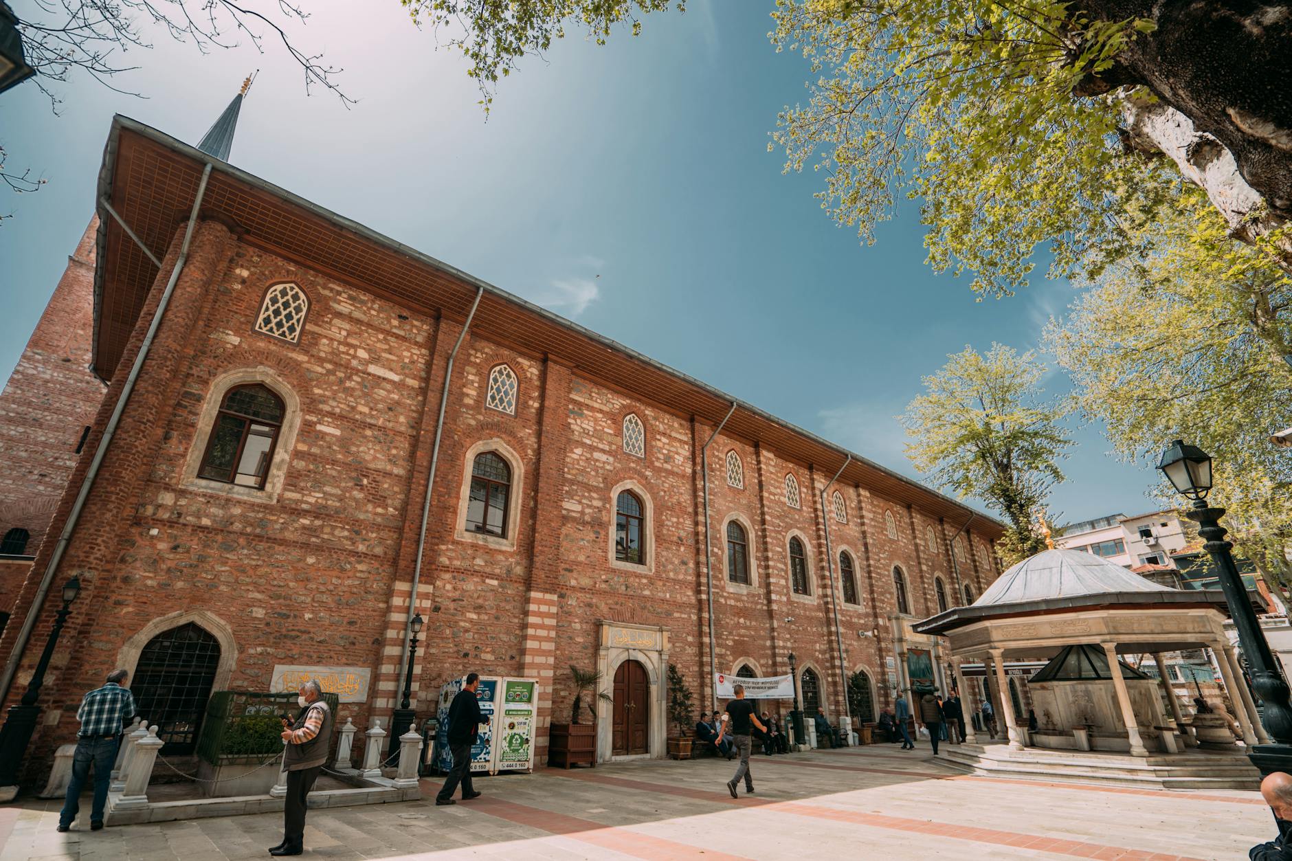A striking, low-angle view of the Arap Mosque's distinctive red brick facade and stone trim, highlighting its historical architecture in Istanbul's Karaköy district. This structure embodies the unique charm discussed in 'A Gothic Secret in the Heart of Karaköy: Why I Love the Unique Arap Mosque,' featuring arched windows, a small minaret peak visible above, and a traditional public fountain in the foreground under a bright blue sky and lush green trees.