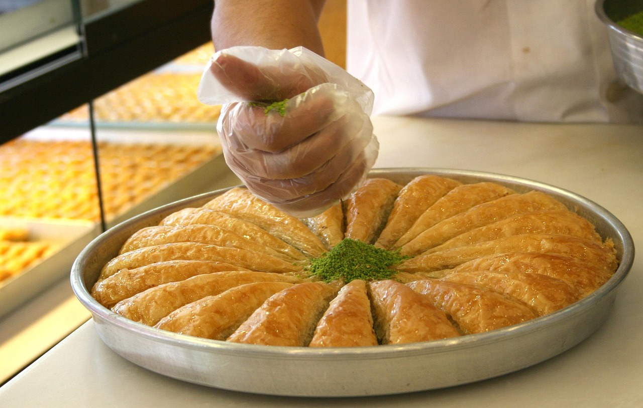 A baker sprinkles finely ground green pistachios over a freshly baked tray of baklava.