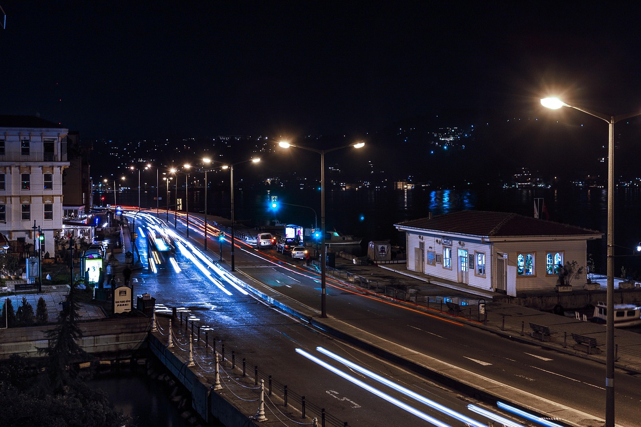 A dramatic, long-exposure photograph capturing the vibrant nightlife along the Bosphorus coast, likely near Bebek or Arnavutköy, illustrating the intersection of urban life and the sea. Streaks of white and red light from moving vehicles blur along the wet road beside the water, contrasting with the dark night sky and the brightly lit windows of a small, traditional white waterfront building. This scene perfectly evokes the ambiance discussed in "Where the City Meets the Sea: My Practical Guide to the Arnavutköy and Bebek Coastline."