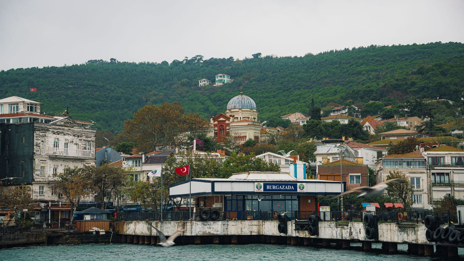 Burgazada ferry terminal sits beneath green hills and a historic church dome.