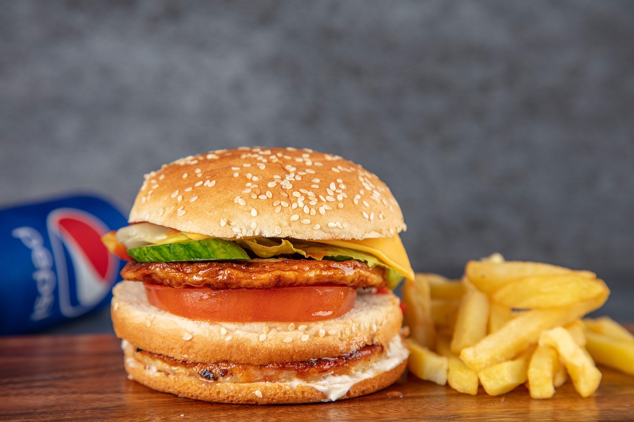 A close-up, professionally lit photo of a sesame seed bun burger, featuring a thick patty, melted cheese, a large slice of tomato, cucumber pickles, and lettuce, sitting on a wooden surface next to a pile of golden french fries and a blurred Pepsi can. While this isn't the specific Taksim wet burger, it captures the general fast-food atmosphere associated with street food favorites, reflecting the spirit of 'I don't care how gross it looks I still love the Taksim wet burger'.