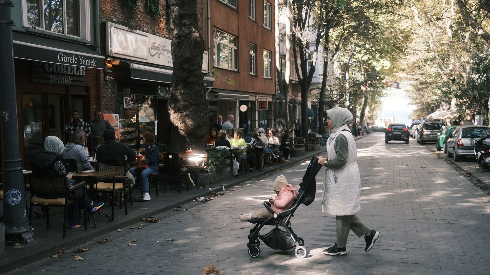 A mother pushes a stroller along a flat pedestrian street lined with trees and cafes.