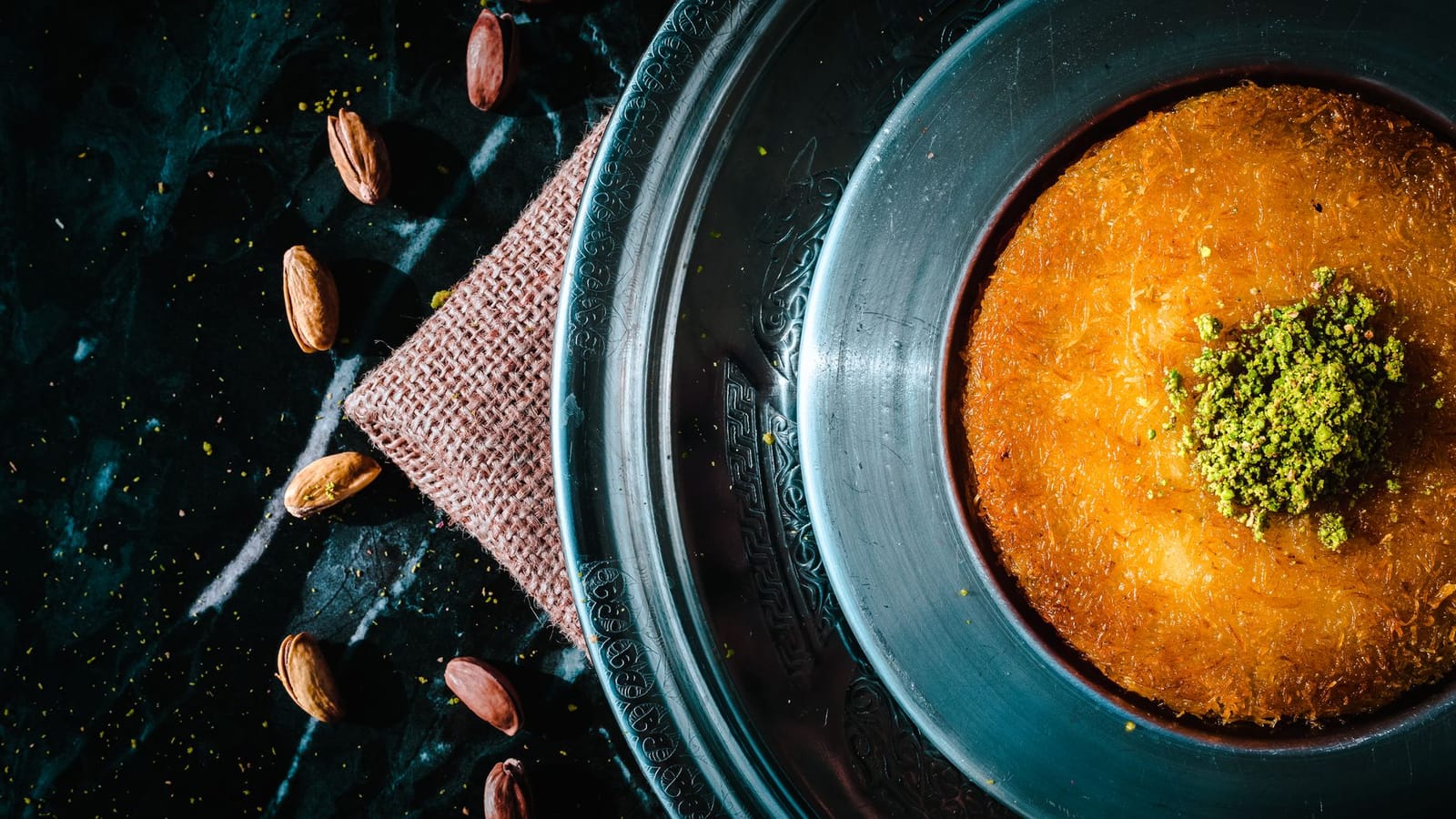 Golden baked kunefe garnished with ground pistachios on an ornate metal tray.