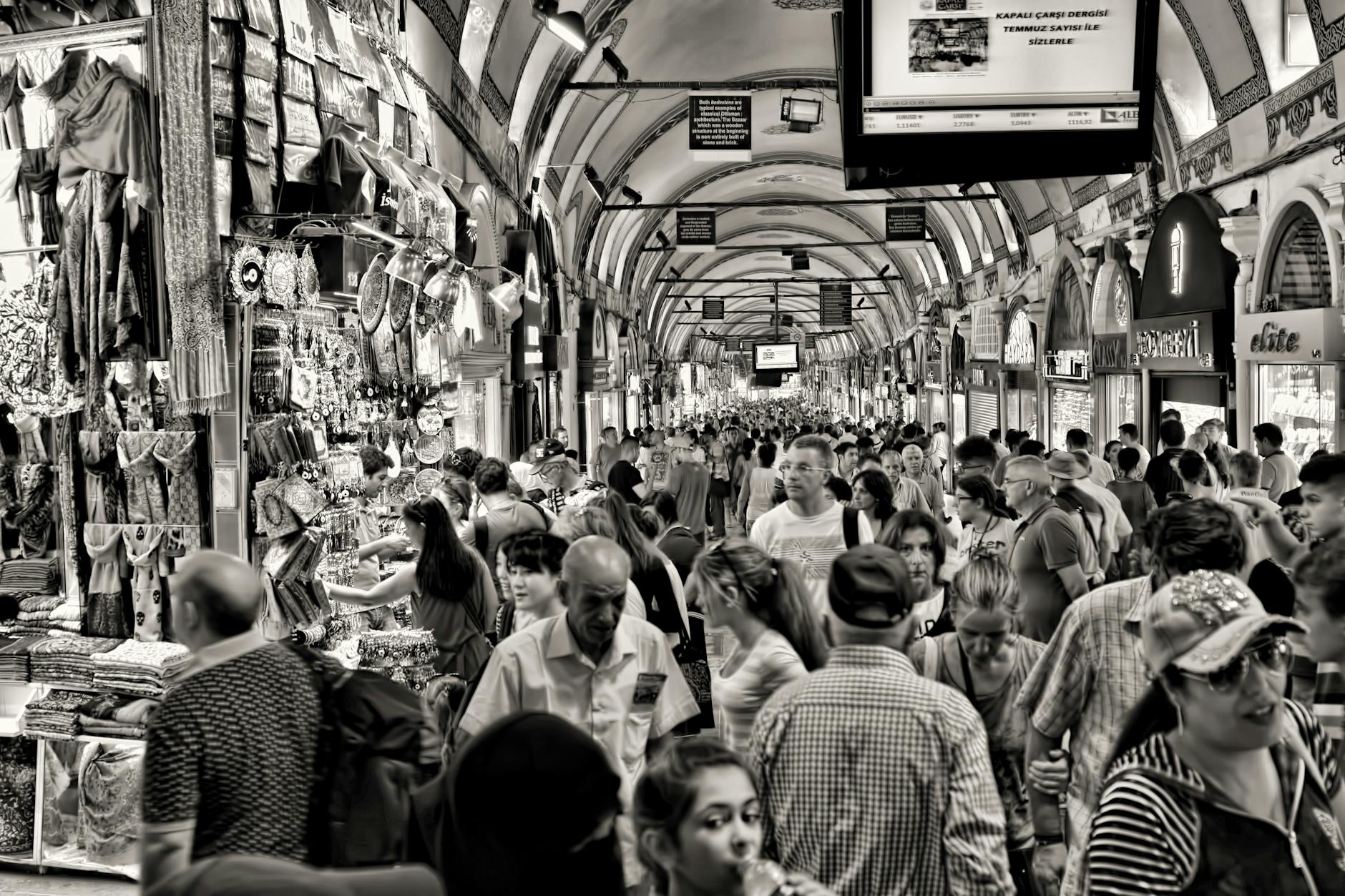 Black and white photo of a crowded interior shopping street within the historic Grand Bazaar in Istanbul, showcasing numerous stalls selling textiles and souvenirs.