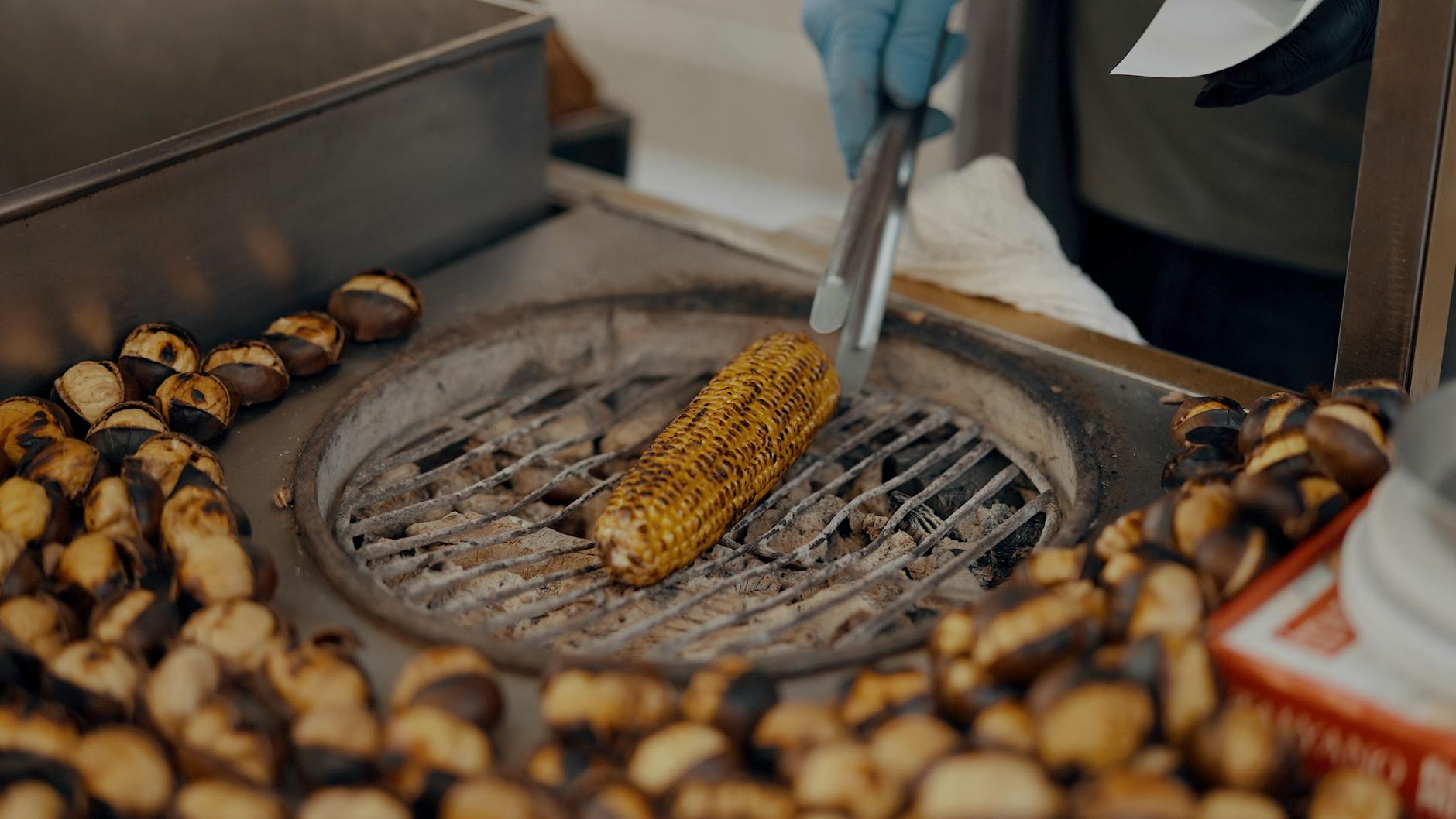 A street food vendor grills corn on the cob alongside roasted chestnuts.