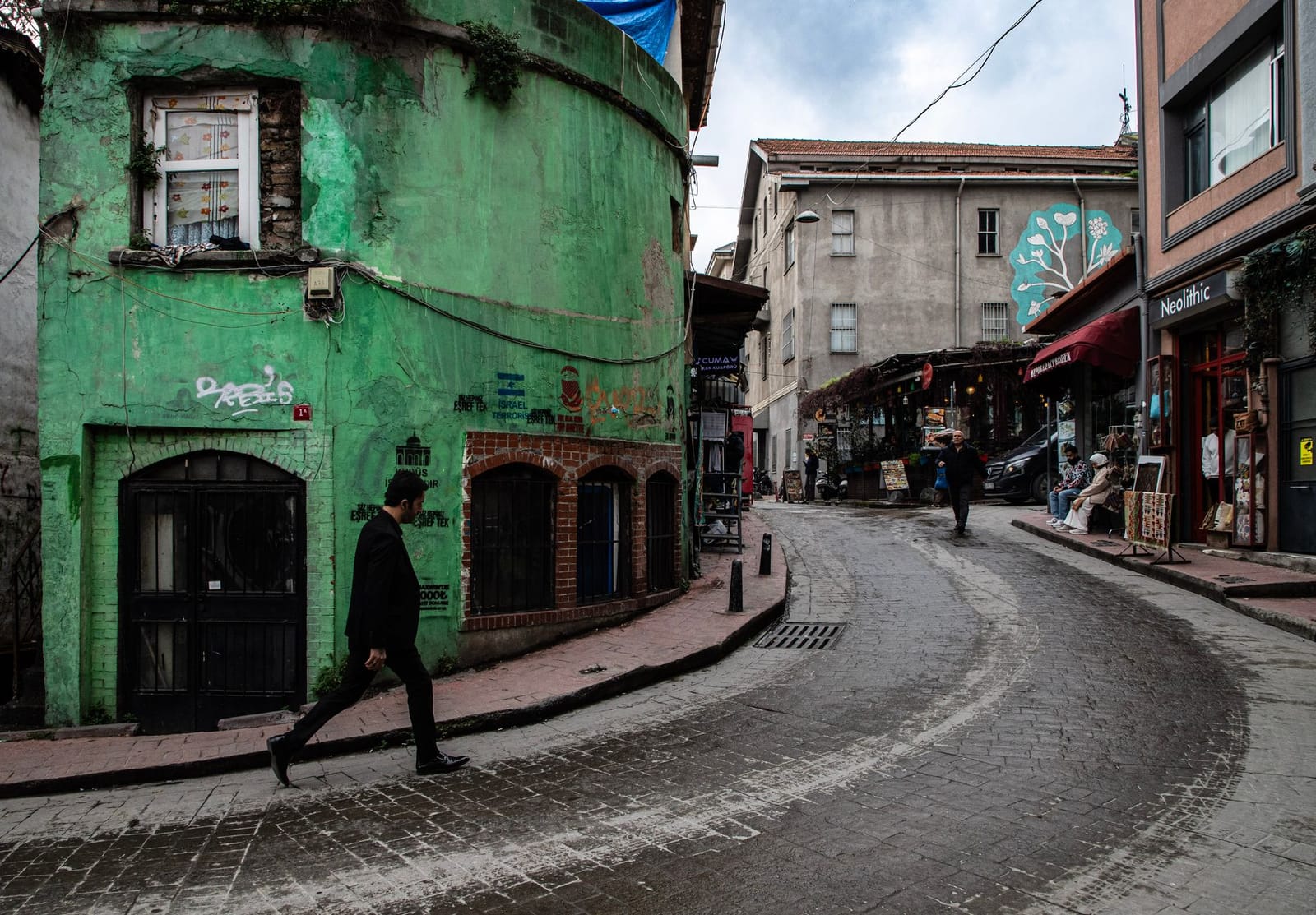 A pedestrian walks down a curving cobblestone street past a bright green historic building.