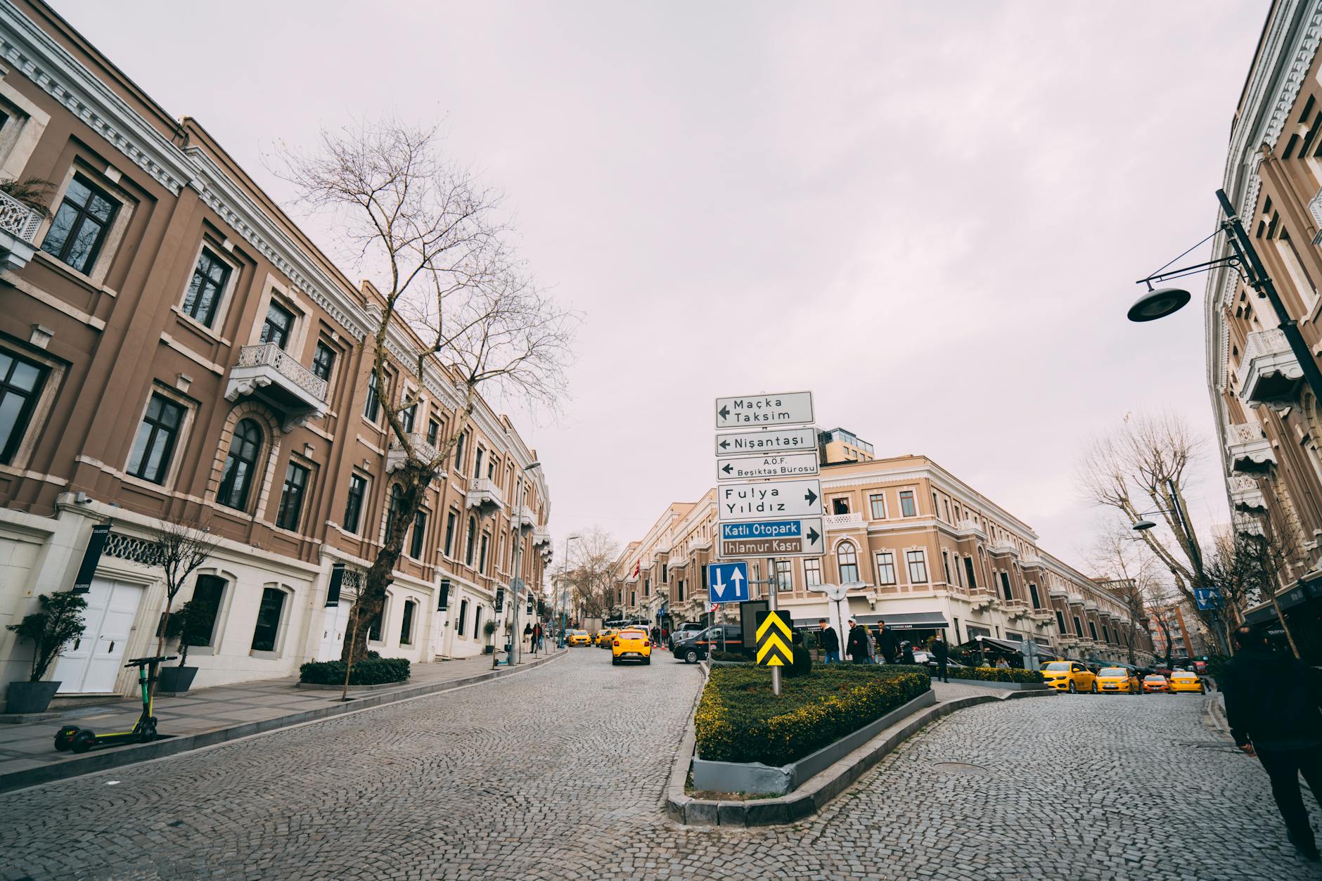 A street scene featuring historic, light brown apartment buildings flanking a cobblestone road in Istanbul. Prominently featured is a traffic signpost directing towards Maçka, Taksim, Nişantaşı, and Fulya Yıldız, indicating the vicinity of Teşvikiye. The presence of yellow Turkish taxis and the distinct architecture suggest this is the area that might be "The Only Reason I Brave the Traffic to Spend an Afternoon in Teşvikiye".