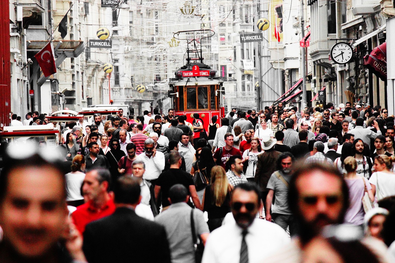 A vibrant, crowded scene on a famous pedestrian street in Istanbul, likely Istiklal Avenue, with a historic red tram (T3 Taksim-Tünel line) visible in the background amidst a sea of people. The image has a slightly desaturated, high-contrast look emphasizing the activity and urban environment, perfectly capturing the lively atmosphere implied by the theme that 'Kurtuluş is loud and the sidewalks are broken but you should stay there anyway'.