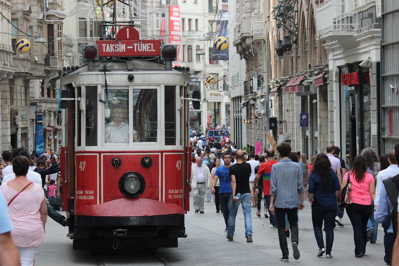 A crowded street scene featuring the iconic red Taksim-Tünel heritage tram numbered 47 moving slowly through a pedestrianized street packed with people, indicative of the lively atmosphere experienced during 'The Grease and Iron of My Favorite Walk Through Perşembe Pazarı' in Istanbul.