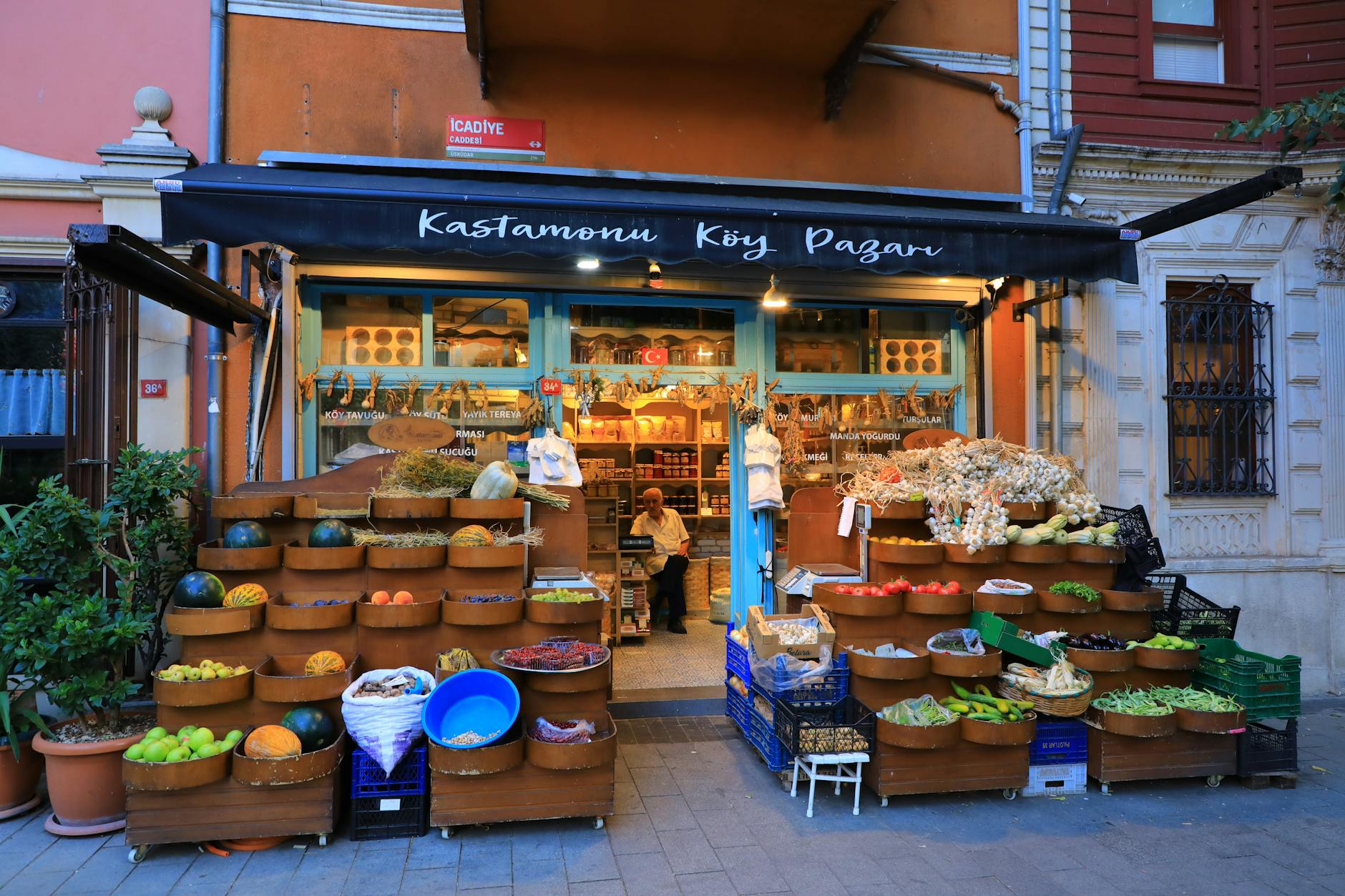A vibrant, traditional Turkish village market stall, 'Kastamonu Köy Pazarı,' displaying an abundance of fresh produce like melons, apples, tomatoes, and large bunches of garlic outside on tiered wooden stands. This scene evokes the authentic local flavor one might encounter during 'The Grease and Iron of My Favorite Walk Through Perşembe Pazarı' in Istanbul.