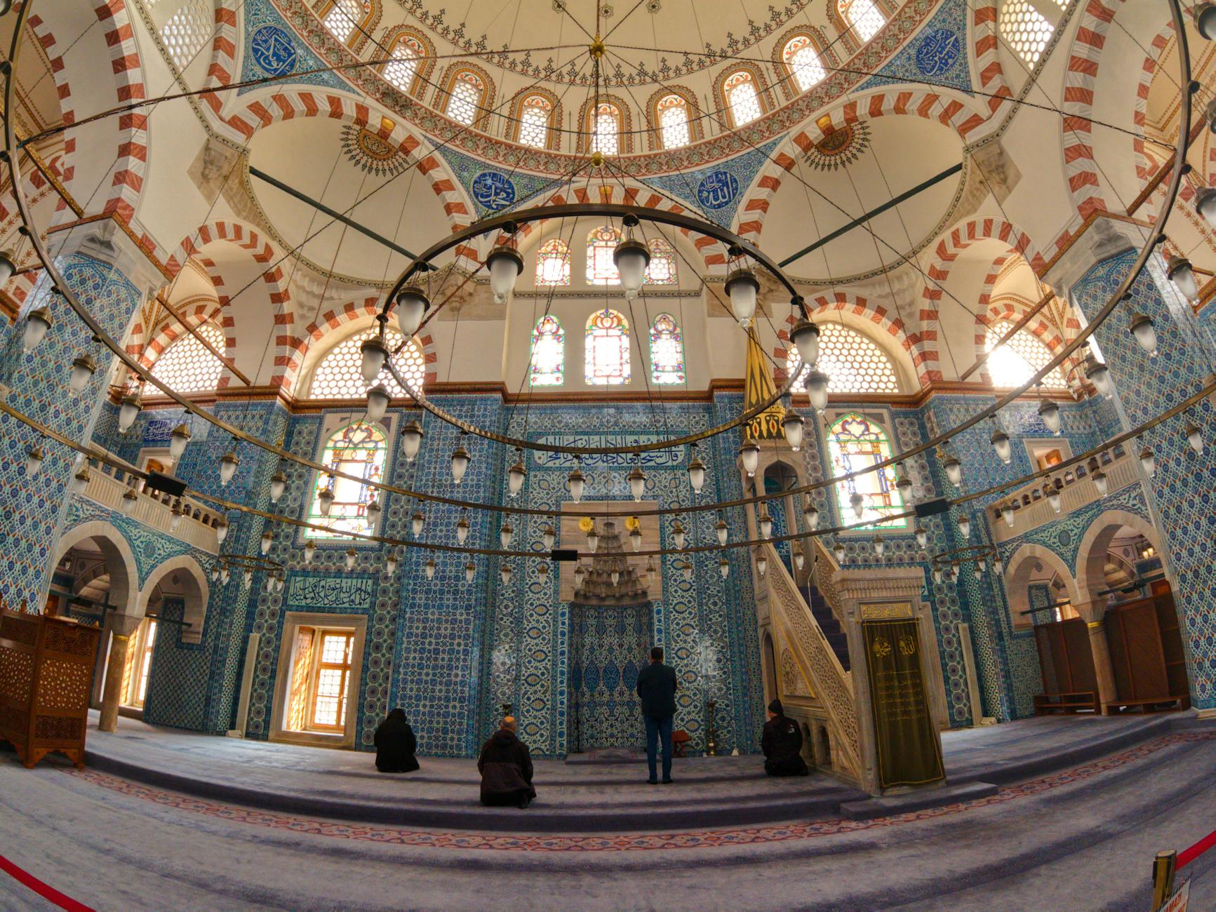 The stunning, brightly lit interior of the Mihrimah Sultan Mosque, showcasing intricate blue Iznik tiling, a vast domed ceiling with patterned arches, and a large circular chandelier. This image perfectly captures the atmosphere described in the article topic: 'A Masterpiece of Light: Why the Mihrimah Sultan Mosque is My Favorite Spot at the City's Highest Point,' highlighting the interplay of natural light entering through numerous windows and illuminating the architectural details.