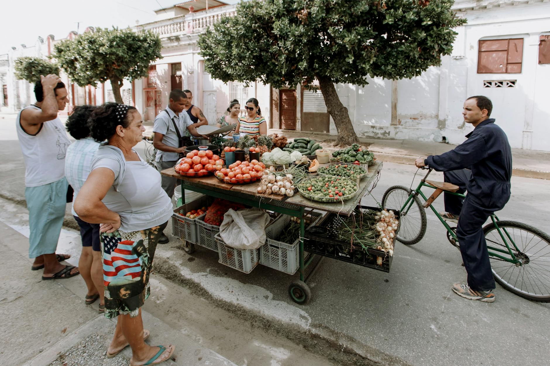 Local residents gather around a mobile produce cart to buy fresh vegetables on a street.