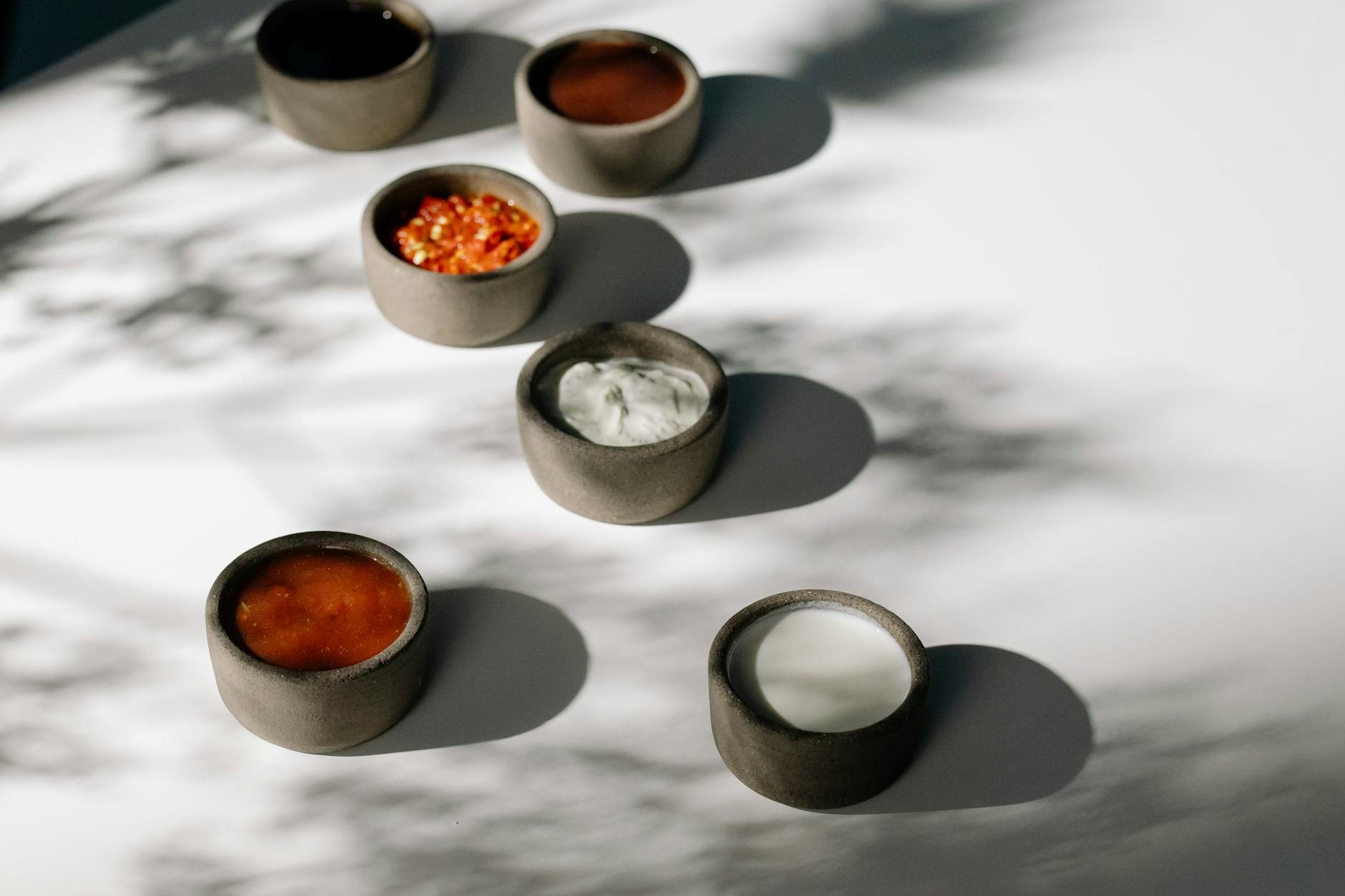 Several small stone bowls containing yogurt and red chili sauces sit on a white surface.