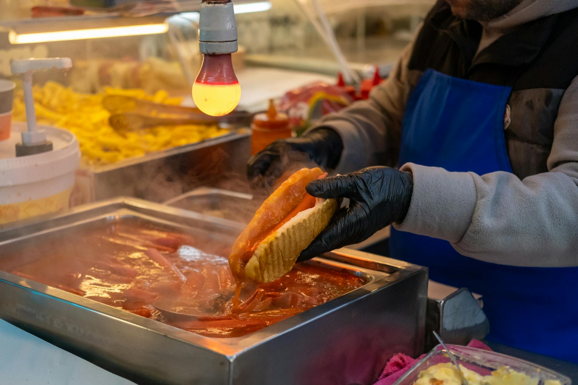 A vendor wearing black gloves and a blue apron is preparing a traditional Turkish wet burger (ıslak burger). The sausage bun is being dipped deeply into a steaming, bright red, savory sauce in a stainless steel container under the warm glow of an overhead light, perfectly illustrating the delicious mess implied by the phrase, "I don't care how gross it looks I still love the Taksim wet burger."
