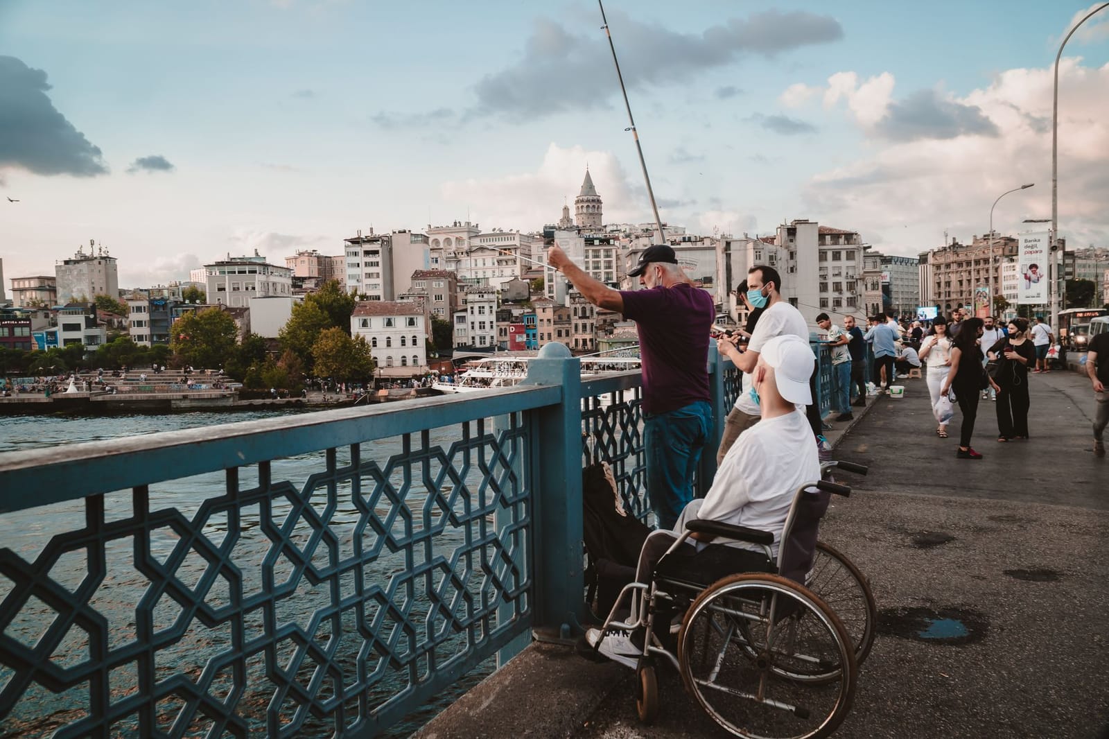 A person in a wheelchair watches fishermen along the flat pedestrian path of Galata Bridge.