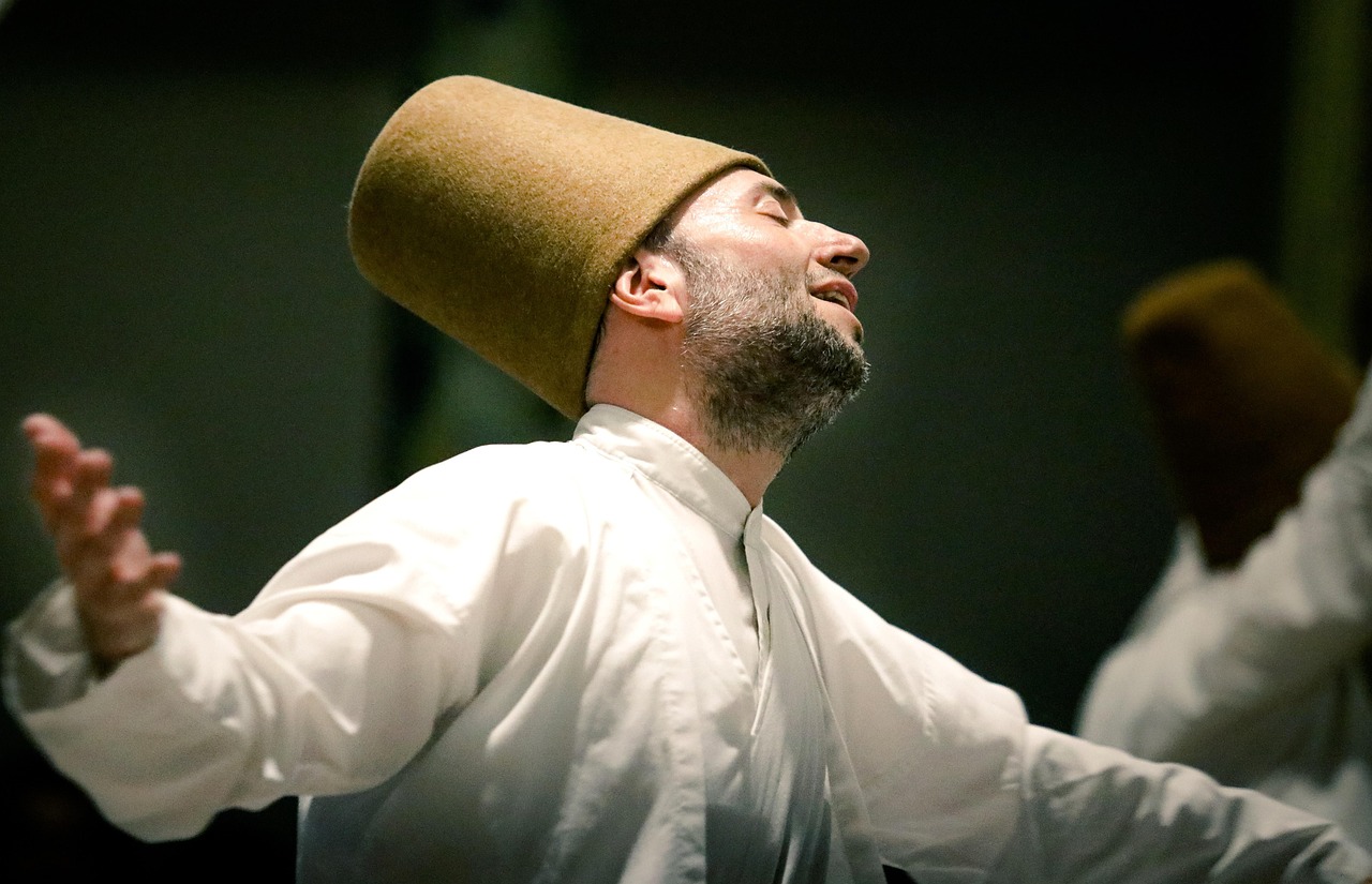 Close-up of a Whirling Dervish (Sema ceremony participant) in a white robe and tall brown felt hat, performing a spiritual dance with eyes closed and arms outstretched.