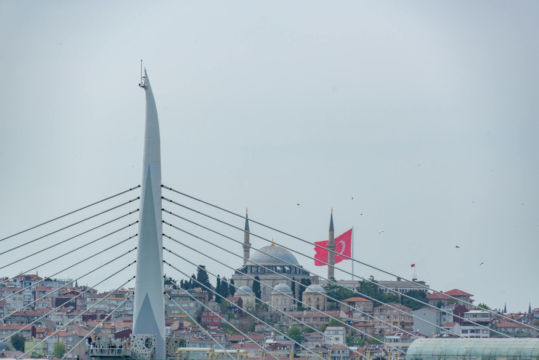Distant view of Yavuz Selim Mosque and a Turkish flag behind bridge cables.