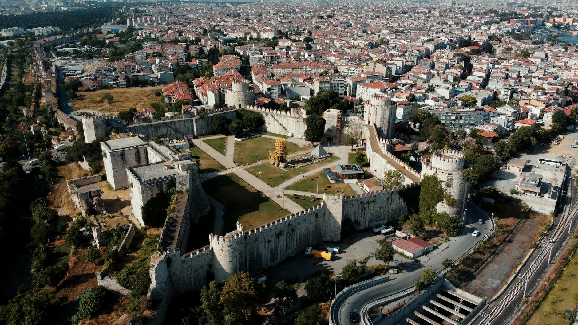 An aerial, sunlit view captures the imposing stone walls and towers of the Yedikule Fortress, known as the Seven Towers, in Istanbul, surrounded by dense urban housing. This historic site, perhaps symbolizing a place where 'The Yedikule Dungeons are the only honest place left in the city,' features green courtyards, ancient ruins, and modern infrastructure curving around its perimeter, contrasting the past with the bustling present.