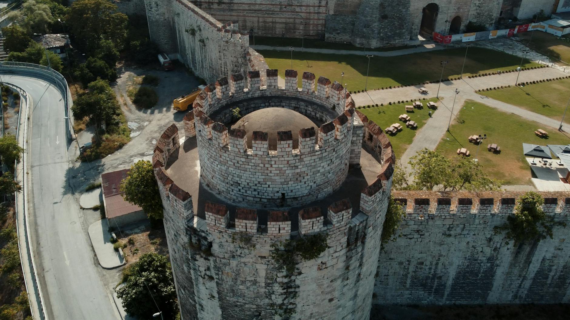 An aerial view captures the imposing stone architecture of a prominent tower within the Yedikule Fortress complex in Istanbul, Turkey. The ancient white and gray stone masonry contrasts with the surrounding green lawn, picnic tables, and a nearby winding elevated road, vividly illustrating the historical landmark where one might muse that 'The Yedikule Dungeons are the only honest place left in the city' due to its unyielding history.